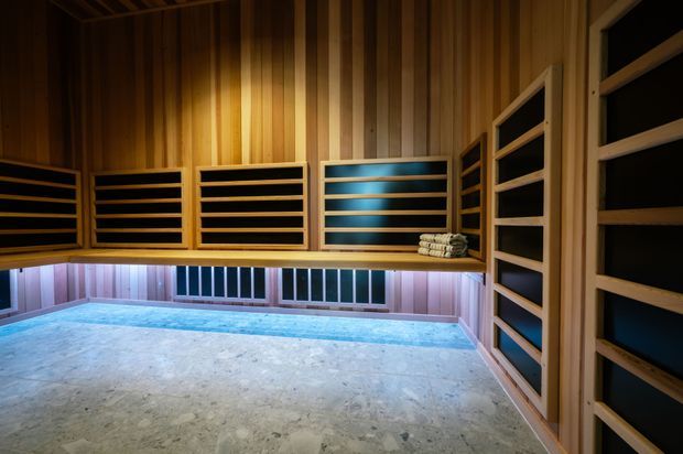 A dimly lit timber-lined sauna with horizontal black-backed ventilation or bench slats, a raised wooden bench, folded towels, and a pale stone or concrete floor. Large lower windows provide cool blue light and contrast with the warm wood interior.