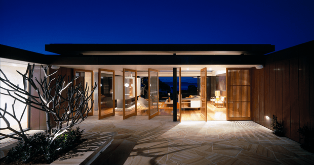 Night-time view of a contemporary single-storey courtyard residence. The open-plan glazed frontage has multiple timber-framed folding or pivot doors opening onto a paved courtyard, with warm interior lighting, lounge furnishings, and a slatted timber screen at the right. A sculptural branching tree and planting occupy the foreground, while dark roof planes and walls frame the composition.