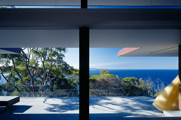 A contemporary open-plan interior or sheltered terrace frames a dramatic ocean view through expansive floor-to-ceiling glazing. Dark structural columns and slim framing contrast with pale tiled flooring, while a projecting pink-toned architectural canopy and a partial yellow upholstered element add colour. Native coastal trees and deep blue sea are visible beyond the terrace.