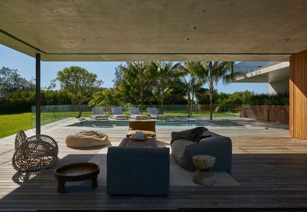 A covered outdoor living terrace with timber decking, a broad timber-lined ceiling, glass balustrades, and relaxed lounge seating overlooking a rectangular swimming pool and landscaped garden. The space connects to a contemporary home with strong horizontal roof forms and timber wall cladding. Bright, resort-like indoor-outdoor setting with tropical planting.