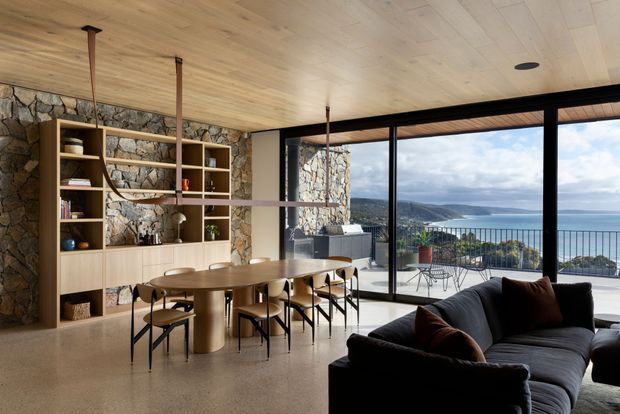 Contemporary open-plan living and dining area with a stone feature wall, integrated timber shelving and cabinetry, a timber dining table, and a dark sectional sofa. Full-height glazing opens to a balcony with expansive coastal and water views. A light timber-lined ceiling, exposed pendant cords, and polished concrete or terrazzo-like flooring create a refined natural-material interior.