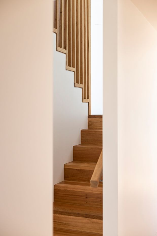 A narrow interior view of a timber staircase ascending between smooth white plaster walls. The stair treads, risers, side handrail, and vertical slatted balustrade are finished in warm natural wood, creating a clean, minimalist architectural detail.