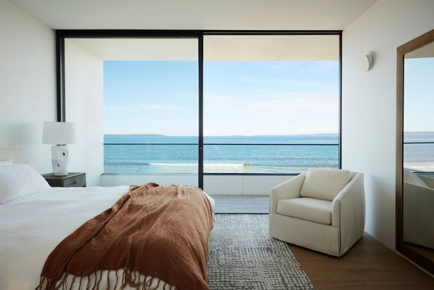 Bright contemporary bedroom with a waterfront outlook through expansive black-framed sliding glazing. The room contains a neatly made bed with a rust-brown throw, a cream upholstered armchair, a small bedside table and lamp, a textured neutral rug, white walls, timber flooring, and a large leaning mirror. A balcony railing and ocean are visible beyond the glazing.