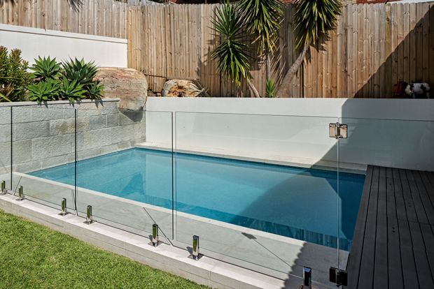 A compact residential swimming pool enclosed by frameless clear glass safety fencing, with a dark timber deck along one side. The pool sits beside a rendered and blockwork boundary wall, with timber privacy fencing, natural stone retaining elements, and established yucca-like planting behind it. Bright outdoor setting with clean contemporary detailing.