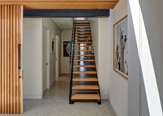 A bright contemporary residential entry hall viewed from the front door, featuring an open-riser timber staircase with slim black metal stringers and handrail. Vertical timber slats frame the entry and continue across the ceiling, while white walls, terrazzo-like flooring, framed artwork, and a dark overhead bulkhead create a clean, warm modern interior.