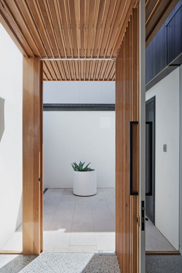 A contemporary residential entryway framed by warm timber slats forming a ceiling canopy and vertical screen. A large glazed timber-framed door with dark hardware opens toward a small, bright tiled courtyard containing a potted succulent. The entry floor is a light terrazzo-like finish, with white walls and dark exterior cladding visible at right.