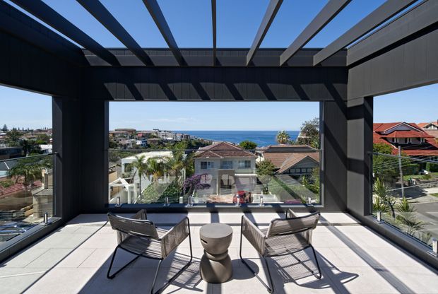 A bright, elevated outdoor terrace or balcony framed by a dramatic dark structural pergola with broad floor-to-ceiling glazing and an open roof grid. Two woven lounge chairs and a small round table face a clear coastal ocean view over a suburban neighbourhood. The space has pale large-format flooring, strong geometric shadows, and a contemporary architectural character.