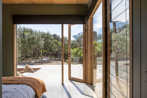 A bright bedroom interior opening onto a timber deck through large timber-framed glazed doors. The room has dark vertical wall panelling, a timber-lined ceiling, pale carpet, and a partially visible bed with a warm brown textured throw. The glazing reflects the interior and frames a native landscaped outdoor setting.