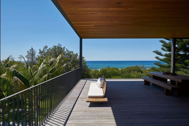 A spacious elevated timber deck overlooking the ocean, framed by a deep covered roof with warm timber soffit and dark slim structural posts. The deck has dark timber flooring, black metal balustrades, a built-in-style bench dining table, and a simple cushioned sun lounger. Tropical vegetation and clear blue water create a relaxed coastal setting.