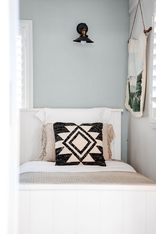 A compact single bed nook with white panelled bed joinery, pale blue-grey walls, white plantation shutters, and a black wall-mounted light. The bed is dressed in white and beige linens with a bold black-and-white geometric cushion, while a fabric wall hanging adds a relaxed decorative touch.