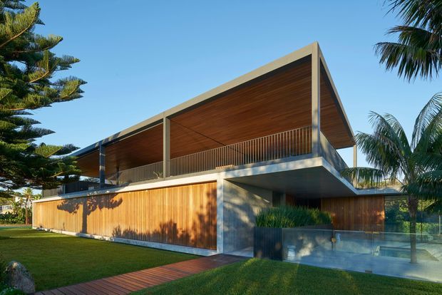 Exterior view of a contemporary two-storey residence with a dramatic pitched roof and deep covered upper-level terrace. Warm timber cladding and soffits contrast with exposed concrete structure, slim dark-framed glazing, and a transparent glass balustrade. The home is set within a landscaped tropical garden with palms and a timber path.