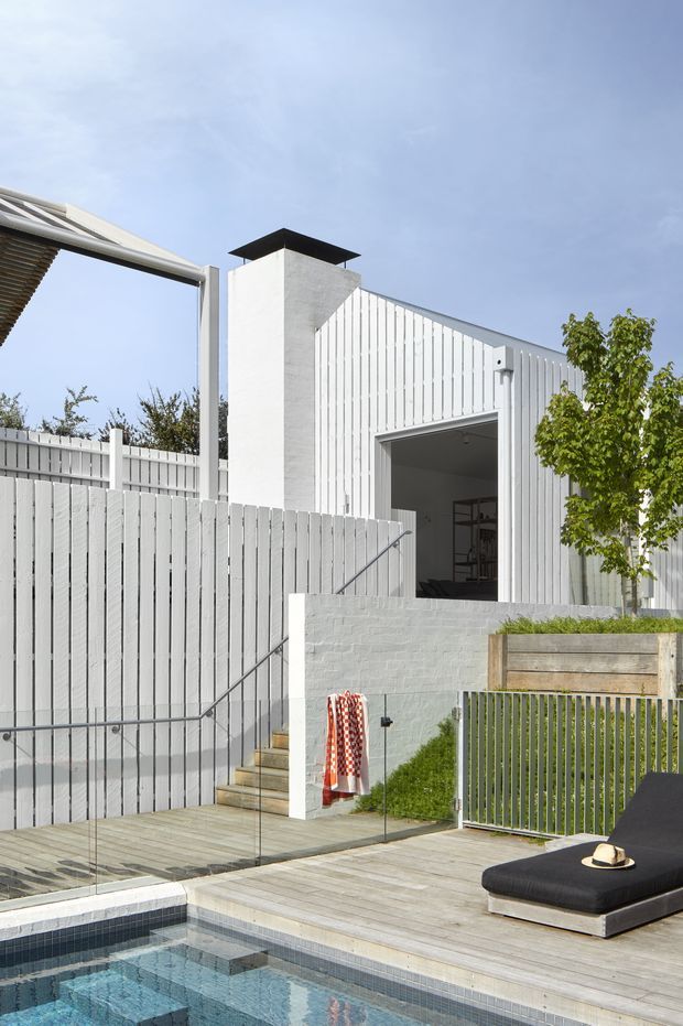 A bright contemporary exterior showing a compact pool courtyard and raised timber deck in front of a white-painted, vertically clad building. Glass pool fencing, pale timber decking, a concrete-block retaining wall, outdoor stairs, metal handrails, and a dark outdoor sun lounger are visible. The building has a simple shed-like roof form with a prominent white rectangular chimney or service tower and a large open doorway.