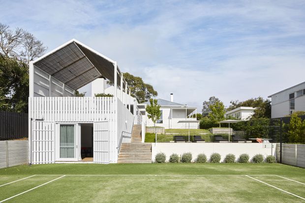 Exterior view of a modern multi-level residential property bordering a small artificial-turf sports court. The white structure features a steep open gable-like roof canopy with exposed slatted detailing, vertical screening, balconies, broad glazing, and integrated stairs leading up to a landscaped terrace. The surrounding architecture is clean-lined and contemporary, with white and grey finishes, dark fencing, and clipped planting.