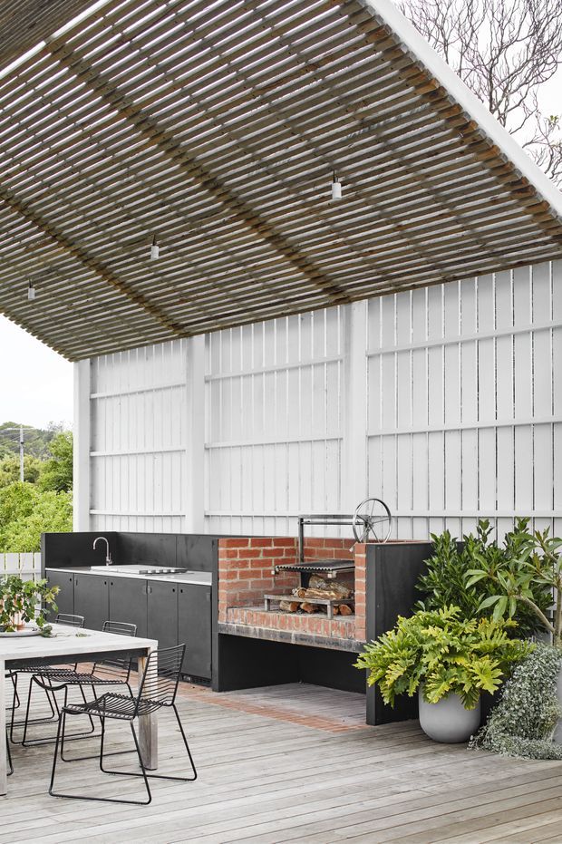 An elevated outdoor entertaining area with a timber-decked floor, white vertical-panel walls, and a slatted timber pergola overhead. A built-in black outdoor kitchen includes a sink and cabinetry, alongside a red brick barbecue or pizza oven. A dining table and wire-frame chairs occupy the foreground, with substantial potted greenery around the perimeter.