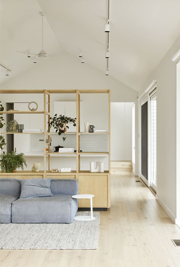 Bright, airy open-plan living room with a high pitched ceiling, pale timber flooring, and a full-height timber open shelving divider separating the sofa area from the adjoining space. The room features a soft blue sectional sofa, neutral rug, white side table, abundant indoor plants, white pendant and track-style lighting, and tall glazed doors along one side. The overall design is calm, minimalist, and Scandinavian-inspired.