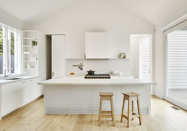 Bright, airy white kitchen with a large central island and two timber stools. The kitchen features white cabinetry, open shelving, a tiled island front, pale timber flooring, integrated cooking appliances, a high pitched ceiling, and generous windows and glazed doors providing abundant natural light.