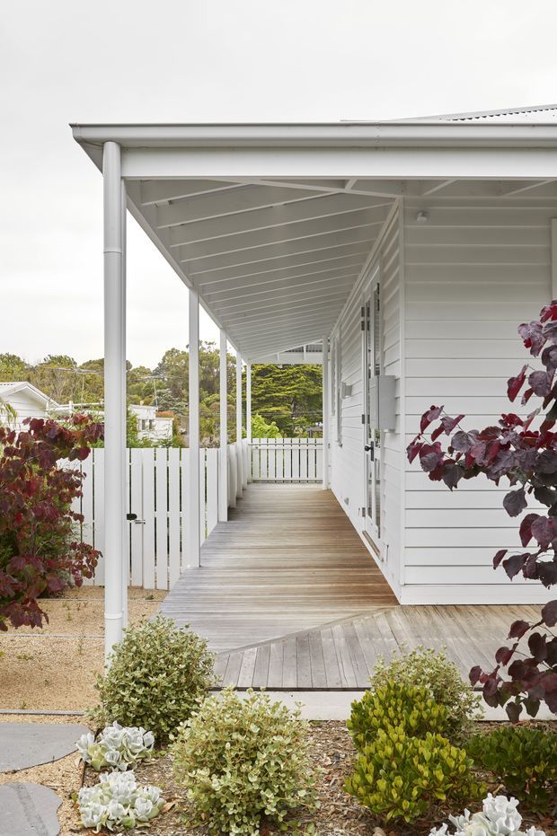 Exterior view of a light-painted home with a long covered verandah and timber decking. The verandah has slender white posts, exposed painted rafters, a white picket-style balustrade, horizontal weatherboard cladding, and doors along the side of the house. A landscaped garden with shrubs and succulents borders the deck, with neighbouring homes and mature trees visible beyond.