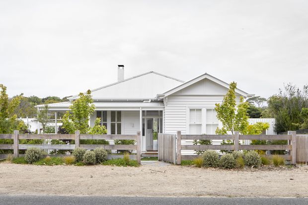 Front exterior view of a single-storey white weatherboard house with a pitched gabled roof, verandah, white-framed windows and doors, and a rustic timber post-and-rail fence. Low coastal-style landscaping and a gravel verge sit between the home and the road.