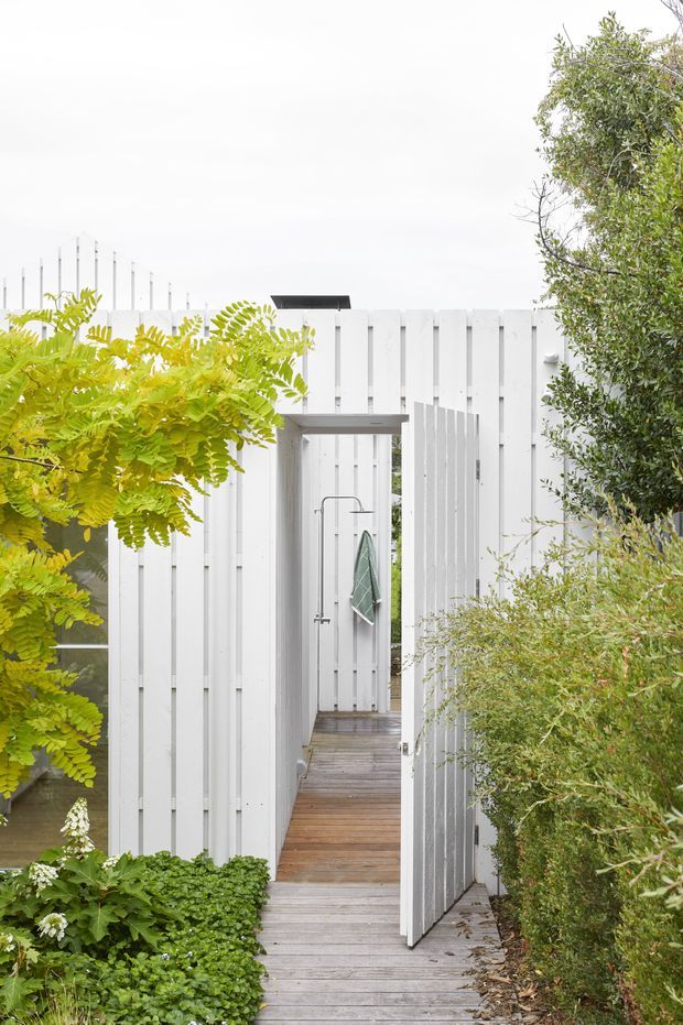 A narrow timber walkway leads through a landscaped garden toward a small white-clad structure or enclosed outdoor area. Vertical white weatherboard-style panels form the surrounding walls and gate, while grey-brown timber decking continues through the passage. Dense green planting frames both sides, and a simple dark projecting canopy marks the entrance.