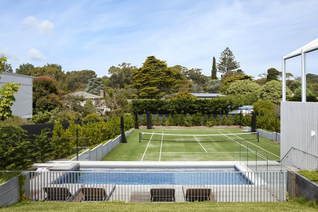 A landscaped residential outdoor recreation area viewed from an elevated position, featuring a full-size fenced tennis court behind a rectangular swimming pool. The scene includes manicured hedges, mature trees, rendered and corrugated metal boundary structures, and a bright open sky.