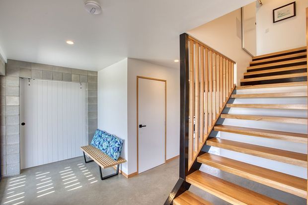Bright contemporary residential entryway featuring an open timber staircase with black structural framing and vertical timber balustrade, a white-panelled storage door set within a grey blockwork surround, and a simple timber bench with patterned cushion. The staircase leads to an upper landing, while recessed ceiling lights and warm timber trims add understated detail.