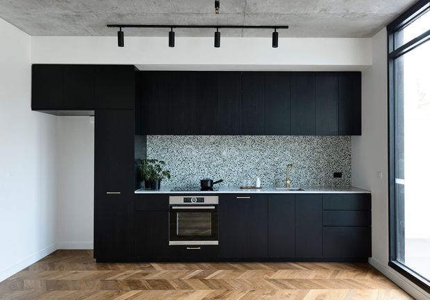 A compact contemporary kitchen with full-height matte black cabinetry, integrated appliances, a speckled terrazzo-style backsplash, and a slim stone-look benchtop. The room has a warm herringbone timber floor, white walls, a raw concrete ceiling, black track lighting, and full-height black-framed glazing along one side. The composition is minimalist and high-contrast, with a small potted plant and limited countertop accessories.