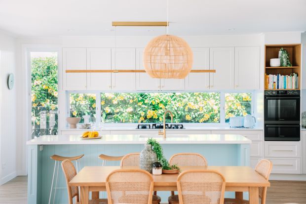Bright, inviting kitchen and dining area with white shaker-style cabinetry, a large pale island, integrated cooktop, timber accents, and a woven pendant light. Floor-to-ceiling windows and a long horizontal window above the benchtop overlook lush greenery. A timber dining table with cane-backed chairs occupies the foreground, while open shelving and built-in ovens are positioned to the right.