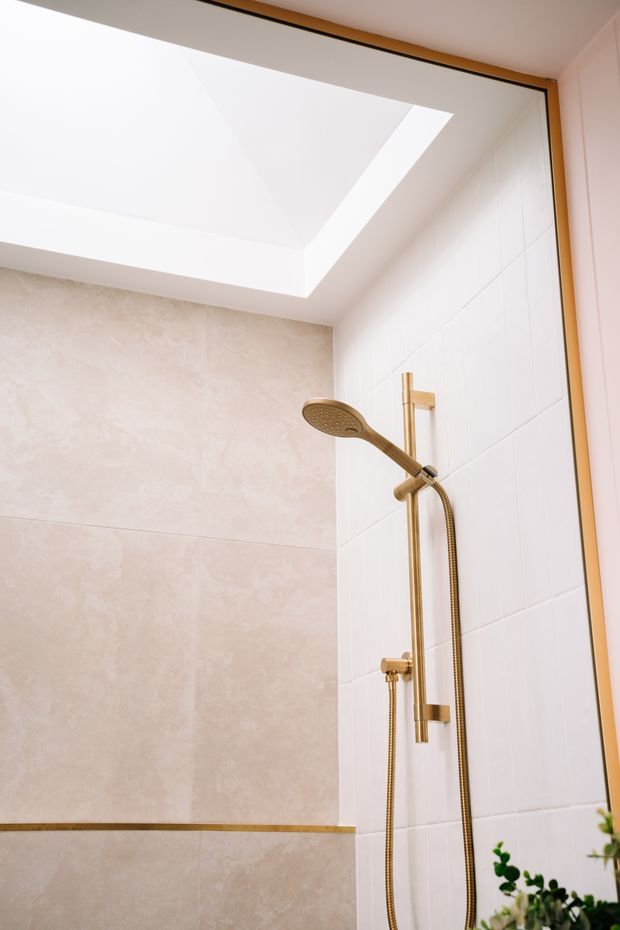 Bright contemporary shower enclosure with a large skylight, warm beige stone-look wall tiles, white rectangular wall tiles, and brushed gold shower hardware and trim. A plant is partially visible at the lower edge.