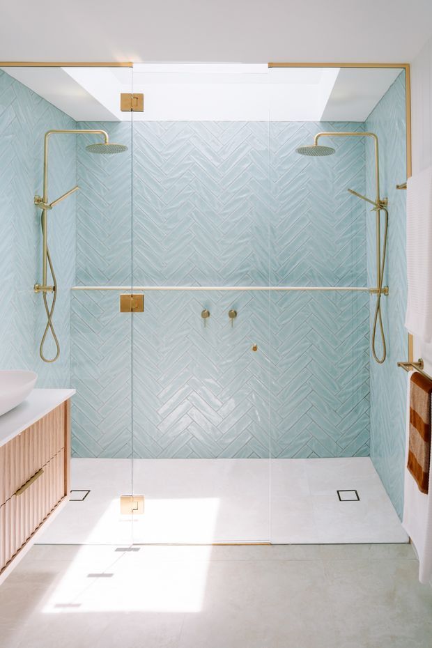 Bright contemporary bathroom featuring a large open walk-in shower enclosed by frameless glass. The shower walls are clad in pale blue-green herringbone tile, complemented by brushed brass/gold shower fixtures, hinges, and trim. A white shower floor, light neutral flooring, and a fluted timber vanity with a white vessel basin are visible at the left. White ceiling and walls create an airy, refined atmosphere.