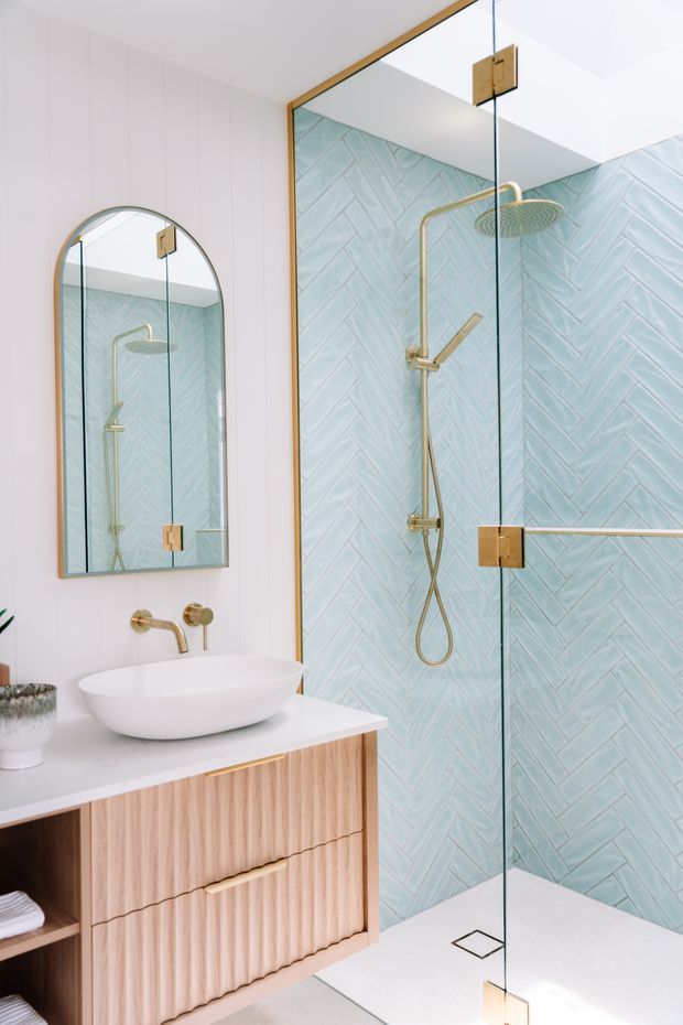 Bright contemporary bathroom featuring a walk-in glass shower with pale aqua herringbone wall tiles and brushed brass fixtures. A white vessel basin sits on a light timber fluted vanity with integrated drawers, paired with an arched wall mirror and white floor and wall finishes.