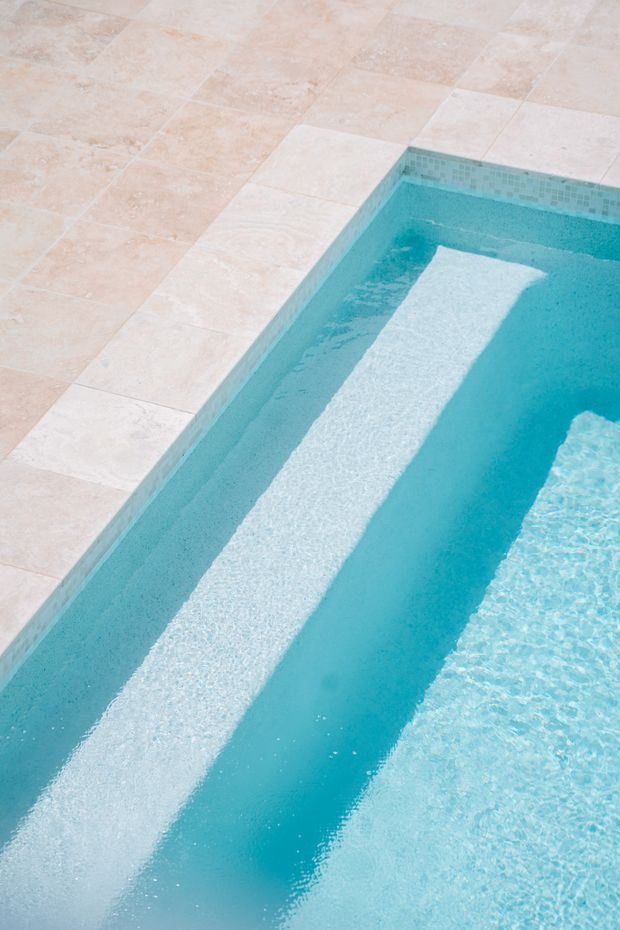 Close-up view of a turquoise swimming pool with a pale stone-tiled coping and surrounding paved terrace. The pool contains a bright submerged or reflected rectangular feature, with rippling water creating a clean, tranquil visual effect.