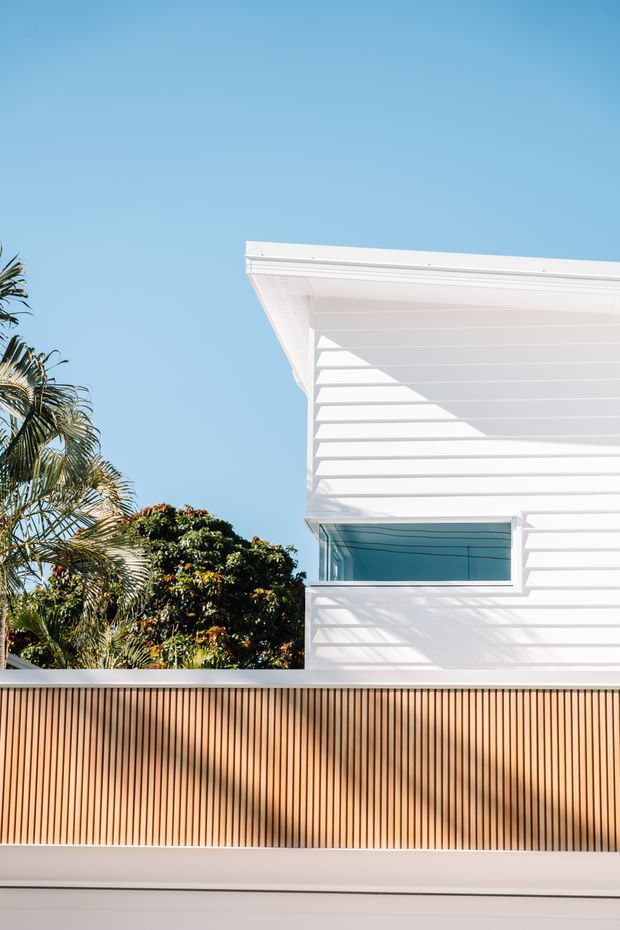 A close-up architectural exterior view of a contemporary white-clad building behind a tall vertical timber privacy fence. The facade features horizontal weatherboard-style cladding, a projecting flat roof edge, and a narrow blue-tinted window. Tropical vegetation is visible to the side, with strong sunlight casting palm shadows across the facade and fence against a clear blue sky.