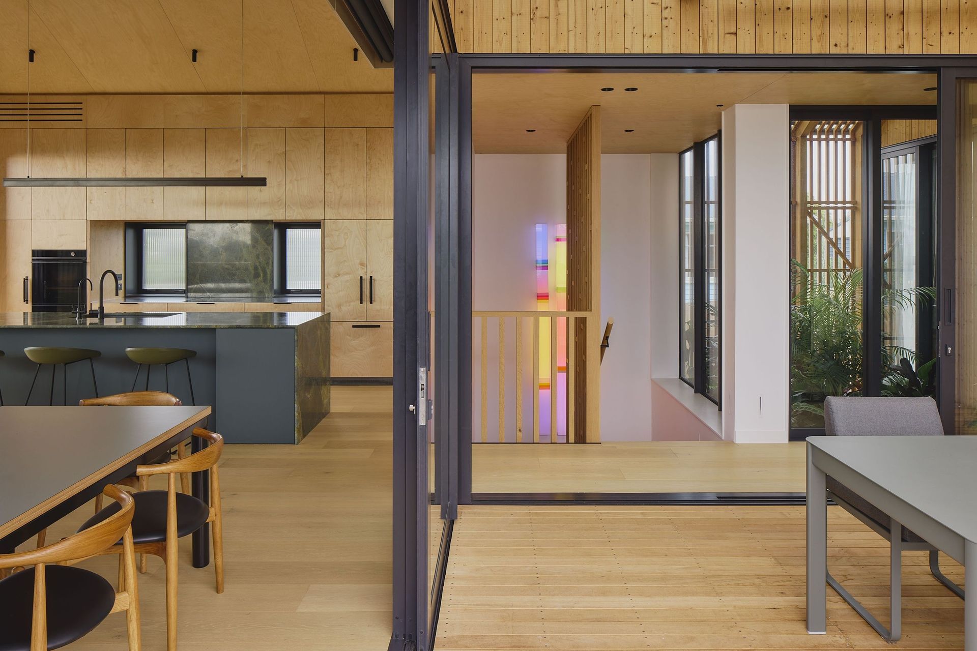 The open-plan living spaces are organised around the internal garden. From the dining room, views extend through the kitchen to the outdoor dining area, staircase and lush courtyard beyond, all unified by plywood ceilings and timber flooring.