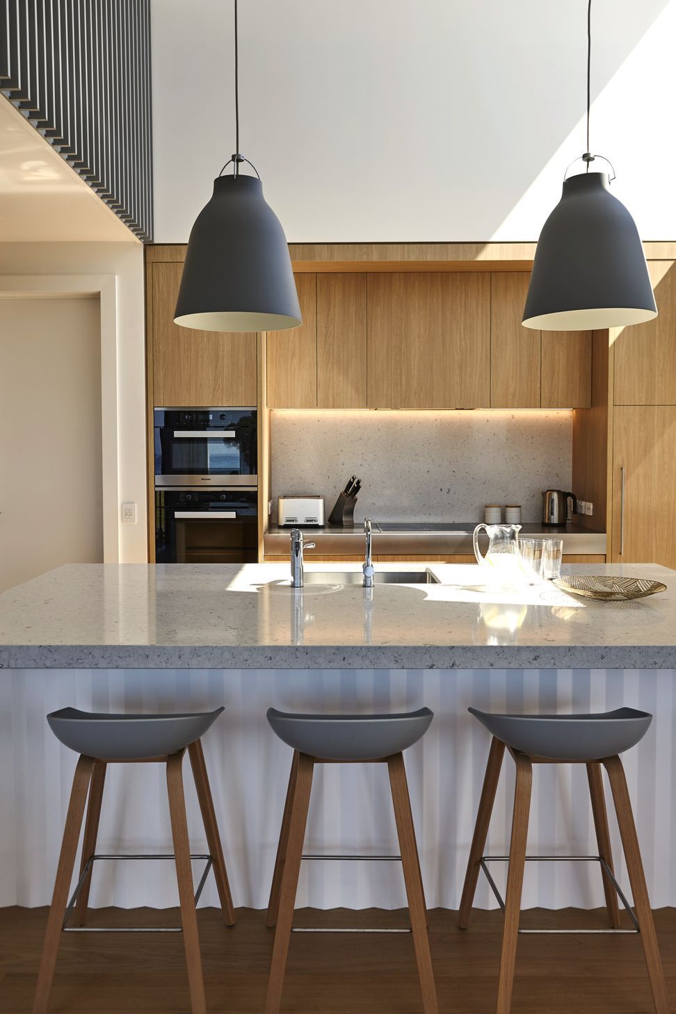 The kitchen is defined by a dramatic double-height ceiling that fills the space with natural light. A generous Caesarstone island sits beneath sculptural pendant lights, while warm oak cabinetry provides a timeless backdrop for family life and entertaining.