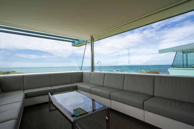 A spacious, minimalist outdoor lounge enclosed by expansive frameless glazing, with a large grey sectional sofa arranged around a reflective rectangular coffee table. The room opens toward a bright ocean and coastal horizon, while a projecting modern roof canopy and slender structural supports reinforce the contemporary architectural character.