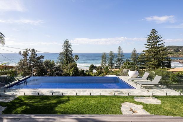 A bright coastal backyard with a rectangular swimming pool overlooking the ocean. The landscaped property includes a manicured lawn, stone paving, glass pool fencing, sculptural white planters, and white loungers, with mature trees and rocky coastal landforms beyond.