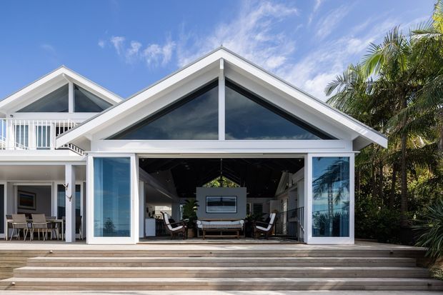 Bright exterior view of a contemporary coastal-style residence with a prominent white triangular gable, extensive dark-framed glazing, and large sliding glass doors opening to a covered outdoor living area. Broad timber decking and stepped terraces lead into the pavilion, which contains lounge seating and a wall-mounted screen. A second gabled volume with balcony railing is visible on the left, while palms and lush planting frame the right side.