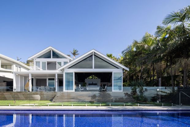 Bright exterior view of a coastal-style multi-level home beside a swimming pool. The facade features prominent white gable roofs, large glazed openings, balconies, white vertical balustrades, and a transparent glass pool fence. Tropical palms and landscaping frame the property.