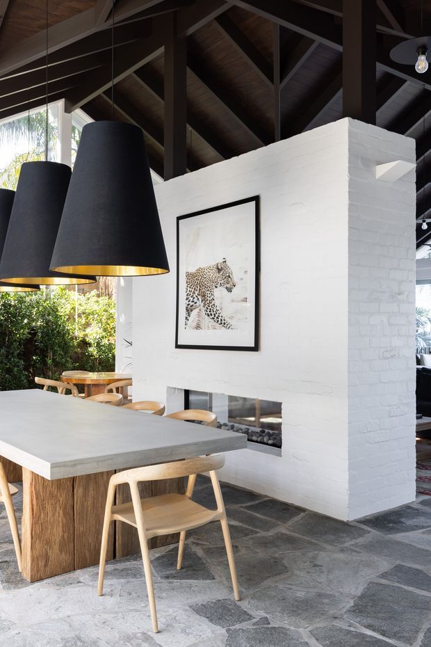A sophisticated open-plan dining area with a large pale stone or concrete-topped dining table, timber base, and light timber chairs. Two oversized black pendant lights with warm metallic interiors hang above the table. A white-painted brick fireplace wall with a framed leopard artwork separates the dining space from an adjacent living area. The room features exposed dark timber roof framing and a dark timber-lined vaulted ceiling, with irregular natural stone flooring and abundant greenery visible through the glazing.