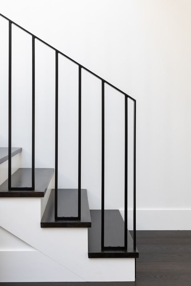 Minimalist interior photograph of a modern stairway with dark timber or stained wood treads, white stair risers and walls, and a slender black metal railing with clear glass infill panels. The staircase descends beside a dark timber floor in a bright, uncluttered space.