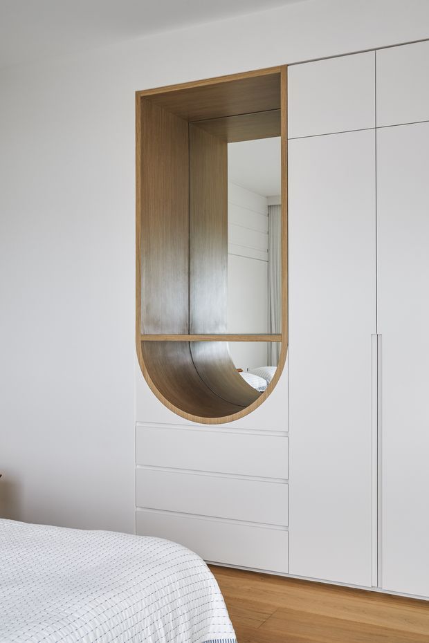A bright, minimalist bedroom with a built-in white wardrobe and integrated drawers. The wardrobe features a distinctive curved, wood-lined open niche with a reflective panel, creating a sculptural dressing or display area. Light timber flooring and the edge of a white quilted bed are visible, with a clean white wall and cabinetry palette.
