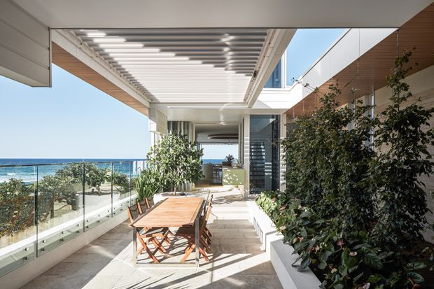 A bright coastal terrace or balcony with a timber outdoor dining table and chairs, bordered by glass balustrades and lush built-in planters. A large louvred pergola and projecting roof structure provide overhead shade, while white and warm timber architectural finishes frame expansive ocean views. The space connects to a modern home through glazed openings.