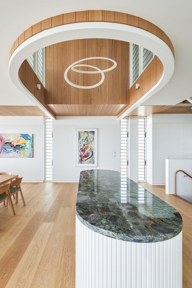 Bright, high-end open-plan kitchen and dining area featuring a large sculptural island with a dark green and grey natural stone benchtop, white vertically ribbed cabinetry, and warm timber flooring. The ceiling has an oversized oval architectural feature with timber lining and integrated circular pendant lighting. White louvre-style screens, timber wall panelling, abstract artwork, and a glimpse of a stair opening complete the refined contemporary interior.