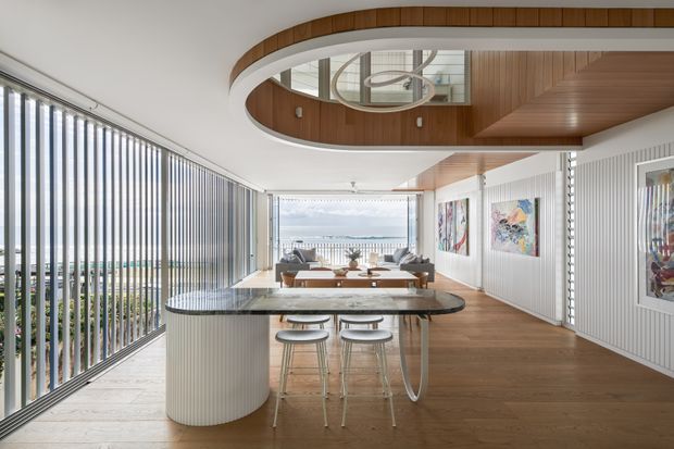 Bright, refined open-plan apartment interior combining a kitchen island with an adjacent living area. The space features warm timber flooring and ceiling panelling, a curved white island with a dark stone benchtop, integrated bar seating, full-height vertical glazing with ocean or coastal views, fluted wall panelling, and a sculptural circular ceiling feature with a contemporary pendant light.