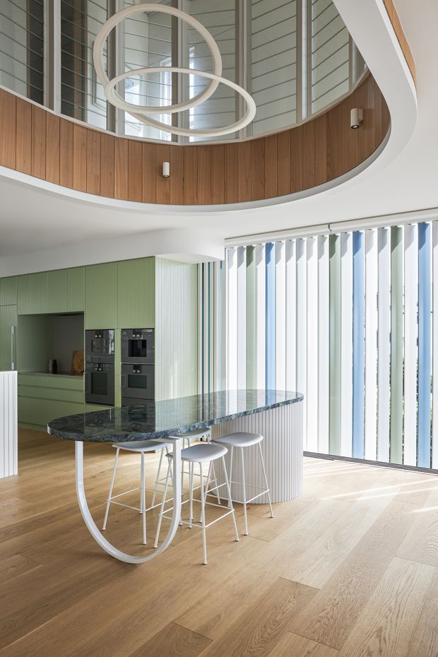 A bright, contemporary kitchen and dining area with a sculptural curved island featuring a dark stone benchtop and integrated rounded support. The kitchen has ribbed sage-green cabinetry, built-in black appliances, pale timber flooring, and a curved white fluted island base. Floor-to-ceiling vertical glazing with alternating fins floods the space with light. Above, a sweeping curved mezzanine or upper-level void is lined with warm timber panelling and framed by large windows, with a modern ring pendant light suspended in the centre.