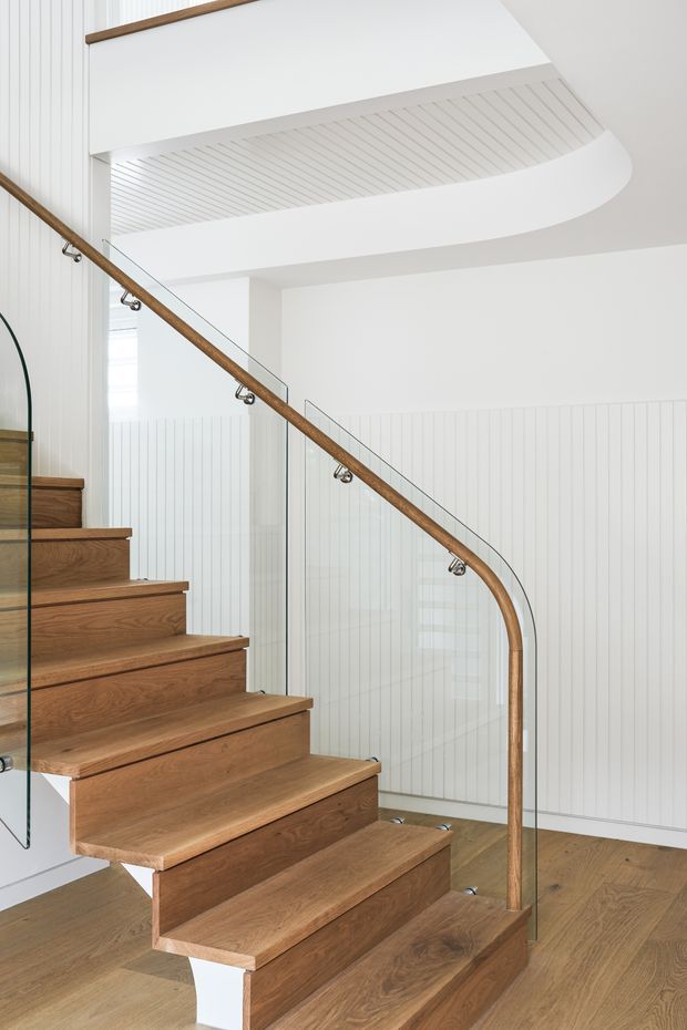 Bright interior view of a contemporary stairway with floating warm timber treads, a clear glass balustrade, and a rounded timber handrail with exposed metal fixings. White vertical wall panelling and a sculptural curved ceiling feature create a clean, refined architectural setting.