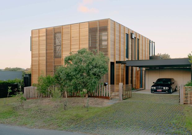Exterior view of a contemporary two-storey residence with a strongly articulated timber screen facade, vertical slatted cladding, and large areas of horizontal timber louvres. A dark flat-roofed carport projects from the house, with a paved driveway and landscaped front yard. The architecture has a warm timber-and-black palette and a highly structured rectilinear form.