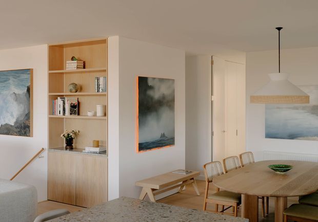 Bright, warm-toned open-plan dining and living area with light timber built-in shelving and cabinetry, a large timber dining table with woven-back chairs, pale upholstered seating, abstract artwork, and a sculptural white pendant light. A stair railing and hallway are also visible, creating a calm contemporary residential interior.