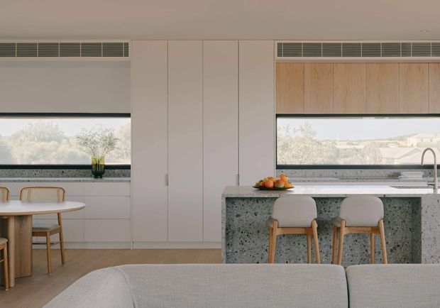 A bright, refined open-plan kitchen and dining area viewed from behind a light upholstered sofa. The space features pale built-in cabinetry, a large terrazzo island with rounded-edge bar seating, timber stools, a light timber dining table and chairs, wide horizontal windows with integrated ventilation grilles, and warm timber window cabinetry. Soft natural light creates a calm, contemporary atmosphere.