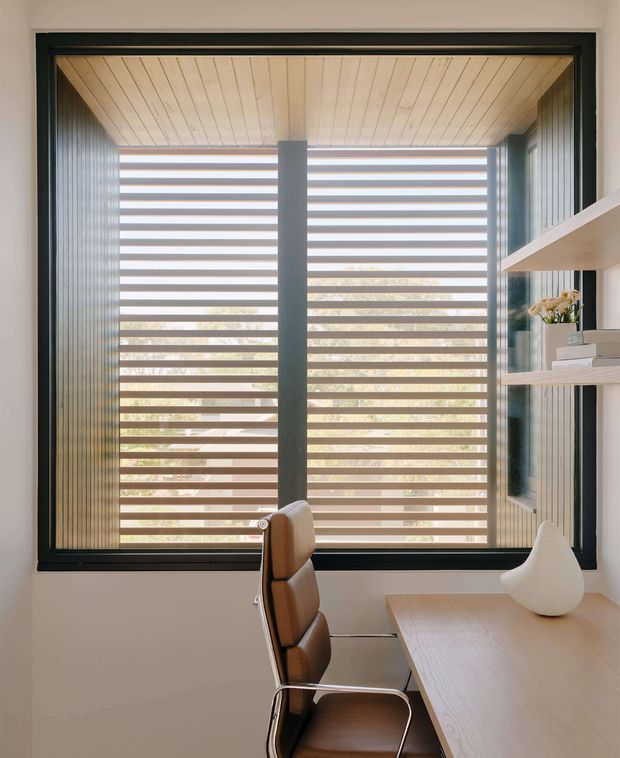 A bright, minimalist workspace positioned beside a large black-framed window. The window features layered horizontal blinds with timber-toned slats, and the surrounding reveal has vertical dark timber panelling. A pale timber desk with a white sculptural vase sits in the foreground, accompanied by a brown leather-and-metal chair. Open pale timber shelving is visible at right, creating a calm, refined home office setting.